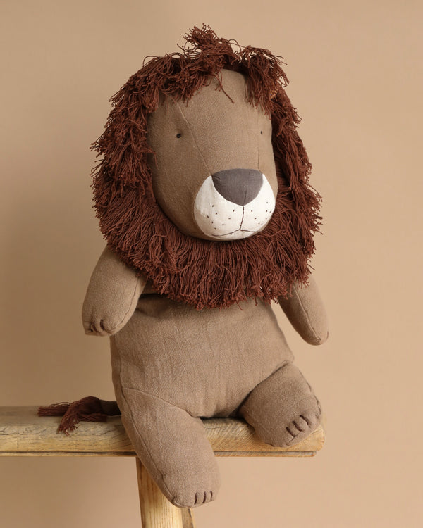 A PRE-ORDER Maileg Safari Friends Big Lion toy, shipping in February, with a shaggy brown mane and a soft, handmade body sits on a wooden bench against a neutral beige background.