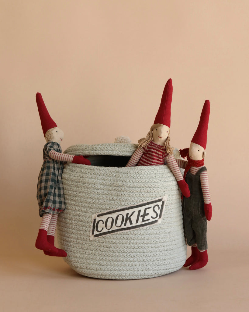 A white woven basket labeled "COOKIES" is encircled by three Maileg Christmas Maxi Pixies dressed in red hats and festive outfits. These delightful cloth dolls, crafted from exclusive materials, add a charming touch to your Christmas decoration on a neutral background.