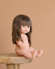 A Minikane Baby Doll (13") - Cherry, with long brown hair and blue eyes, sits nude on a wooden stool against a plain beige background. Clothing sold separately.