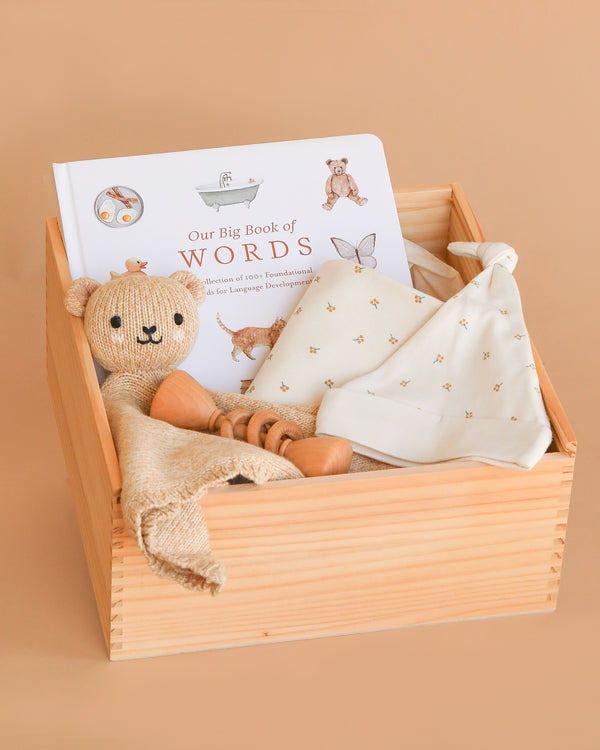 The Newborn Gift Box Set includes "Our Big Book of Words," a knitted bear plush, a printed cream-colored cloth, an organic baby blanket, and a wooden honey dipper, all beautifully arranged in a wooden box.