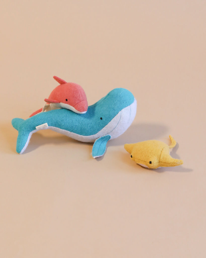 Three colorful Olli Ella Holdie Folk Felt Ocean Animals—a blue whale, a pink dolphin, and a yellow manta ray—on a light beige background.