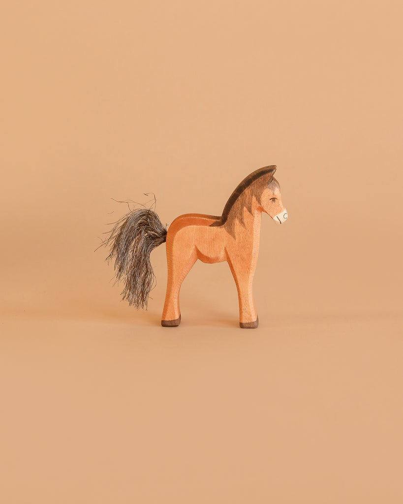 The Ostheimer Brown Colt is a handcrafted wooden horse figurine with a painted mane and tail of dark fibers, standing on a light brown surface—ideal for imaginative play.