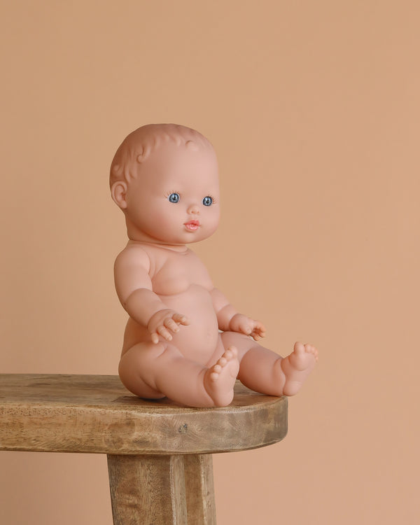 The Minikane Doll (13") - Rosalie sits unclothed on a wooden stool against a beige background. This realistic baby doll is crafted from phthalate-free vinyl for safe play. Clothing sold separately.