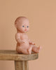 The Minikane Doll (13") - Rosalie sits unclothed on a wooden stool against a beige background. This realistic baby doll is crafted from phthalate-free vinyl for safe play. Clothing sold separately.