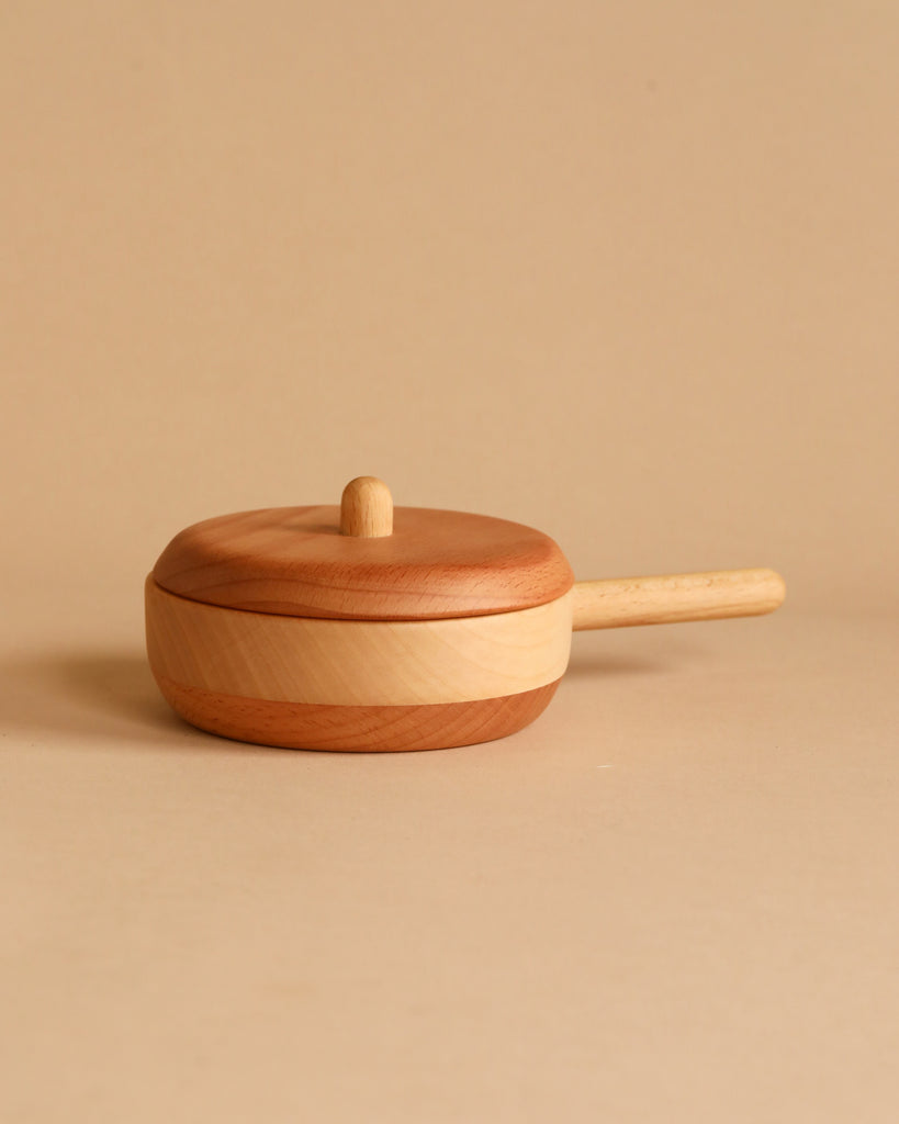 A Handmade Wooden Pretend Pan with a lid and a short handle, crafted from fine hardwoods, is placed against a solid beige background. The pan boasts a natural wood grain finish and is designed in a simple, minimalist style, perfect for timeless pretend play.