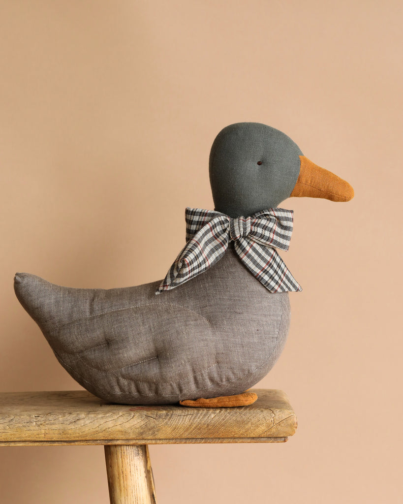 A Maileg Duck Stuffed Animal in gray with a green head, orange beak and feet sits on a wooden bench, adorned with a checkered bow around its neck. It's perfect for festive décor against the soft beige background.