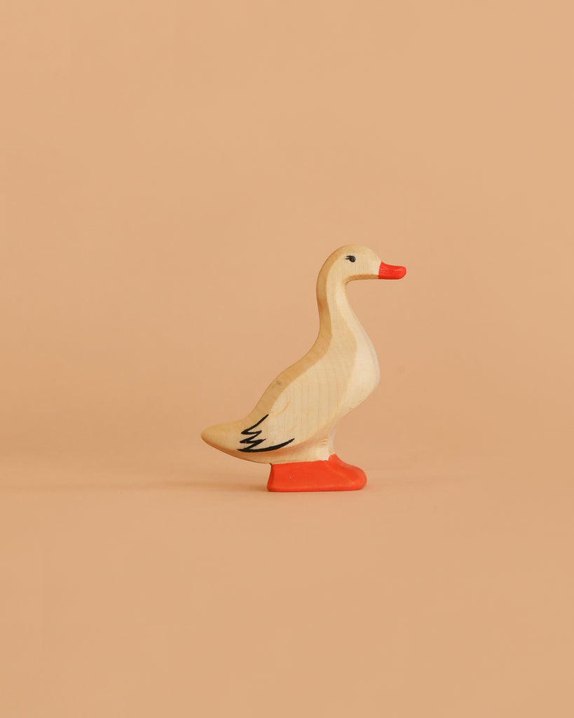 The Bumbu Handmade Wooden Domestic Duck is a small, handcrafted toy featuring a beige body, orange beak, and orange feet, displayed on a solid light peach background.