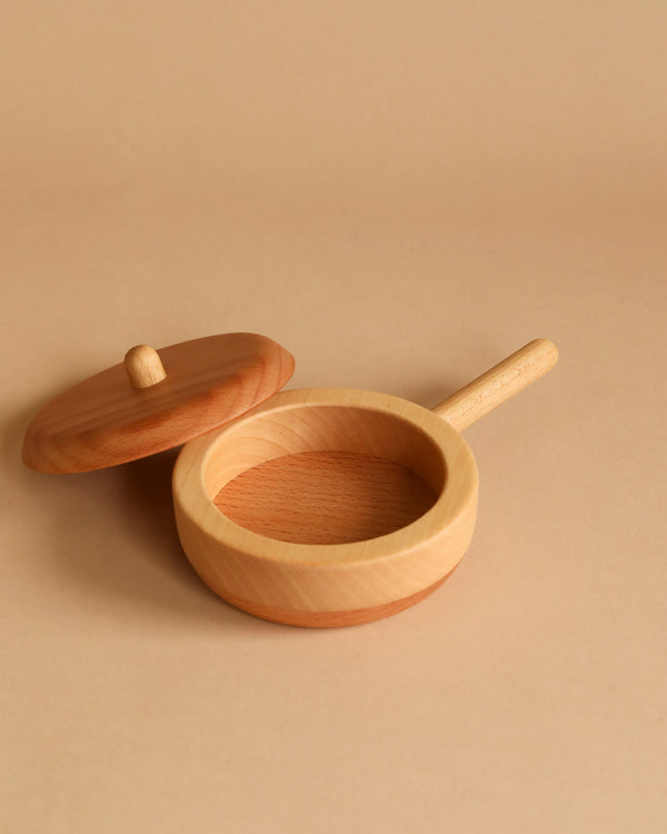 A Handmade Wooden Pretend Pan sits on a beige background. The heirloom-quality pan boasts a smooth, natural wood finish and a handle extending from one side. The round lid, also made of hardwoods, is placed slightly ajar next to the pan, perfect for pretend play.
