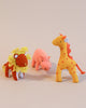Three plush toys from Olli Ella Holdie Folk Felt Savannah Animals—a lion with a mane, a pink pig, and a giraffe with red spots and a bow—stand against a light beige background.