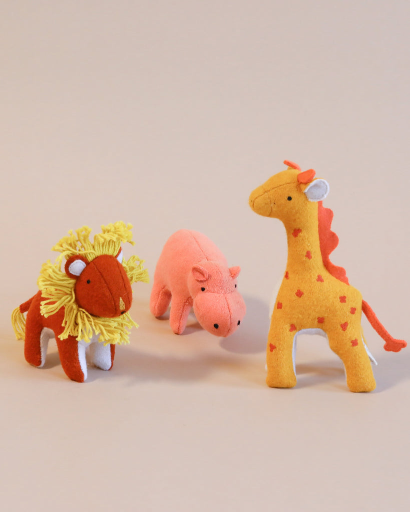 Three plush toys from Olli Ella Holdie Folk Felt Savannah Animals—a lion with a mane, a pink pig, and a giraffe with red spots and a bow—stand against a light beige background.