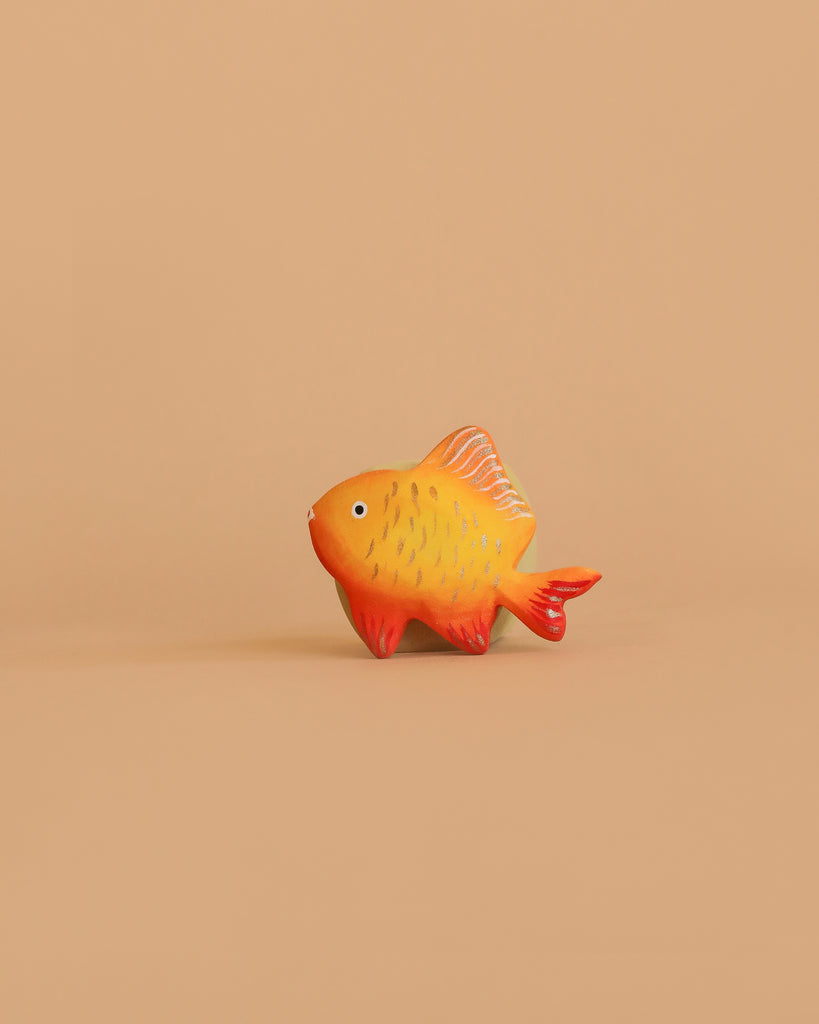 The Bumbu Handmade Wooden Golden Fish, featuring orange and yellow hues with red fins, is displayed against a simple, light brown background.