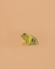 The Bumbu Handmade Wooden Frog, a small green and yellow toy with black spots, is displayed on a plain peach-colored background.