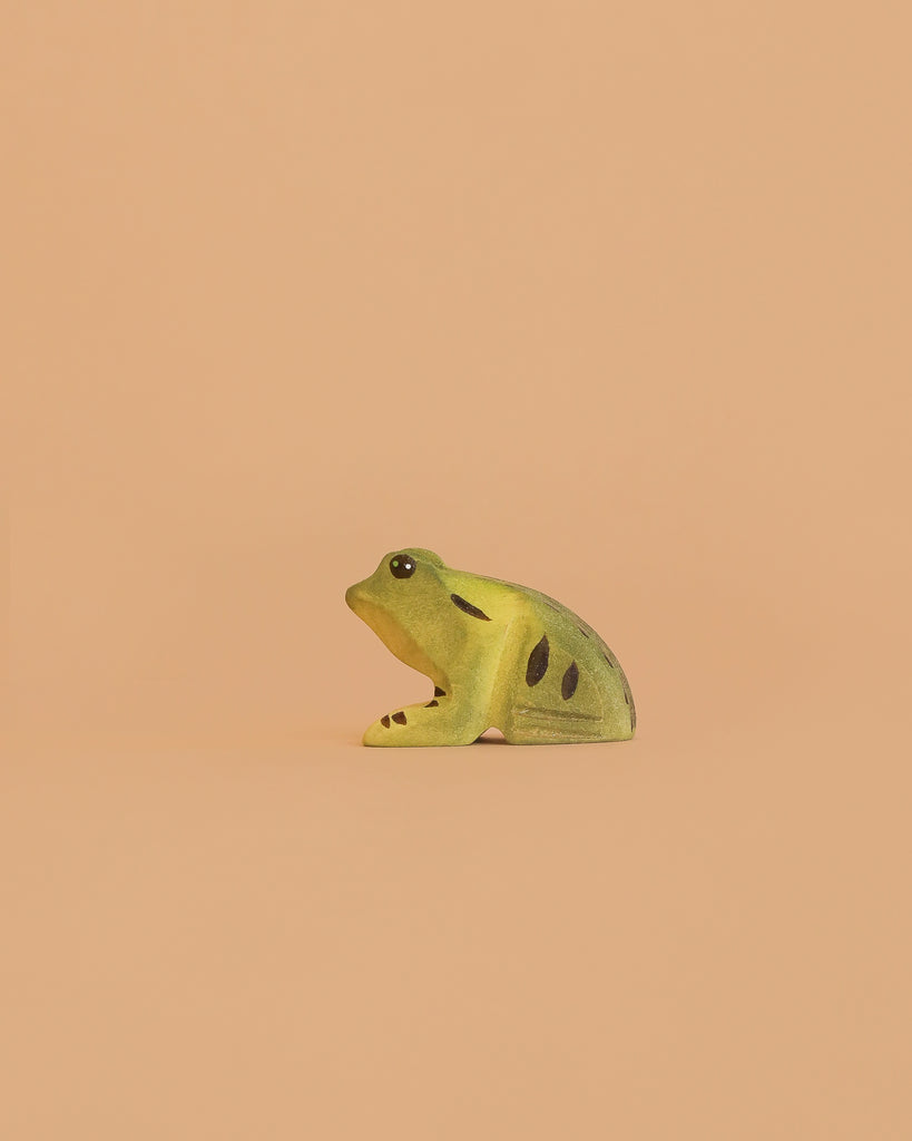 The Bumbu Handmade Wooden Frog, a small green and yellow toy with black spots, is displayed on a plain peach-colored background.