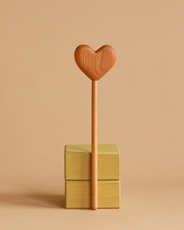 A Handmade Wooden Heart Magic Wand is standing vertically on two stacked golden boxes. The background is a warm beige color, creating a minimalistic and harmonious composition.