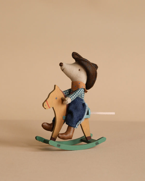The Maileg Cowboy Mouse On Rocking Horse, Little Brother, wears a blue checkered shirt and denim overalls as he sits on a rocking horse with a green base, creating a charming play scene against a beige background.