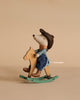 A Maileg On The Go Suitcase Set features a mouse in a cowboy hat, checkered shirt, and blue pants sitting on a wooden rocking horse against a plain beige background.