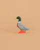 The Bumbu Handmade Wooden Mallard Duck, featuring a green head, yellow beak, gray body, and orange feet, stands on a plain beige background. This charming figurine is crafted from sustainable maple wood.