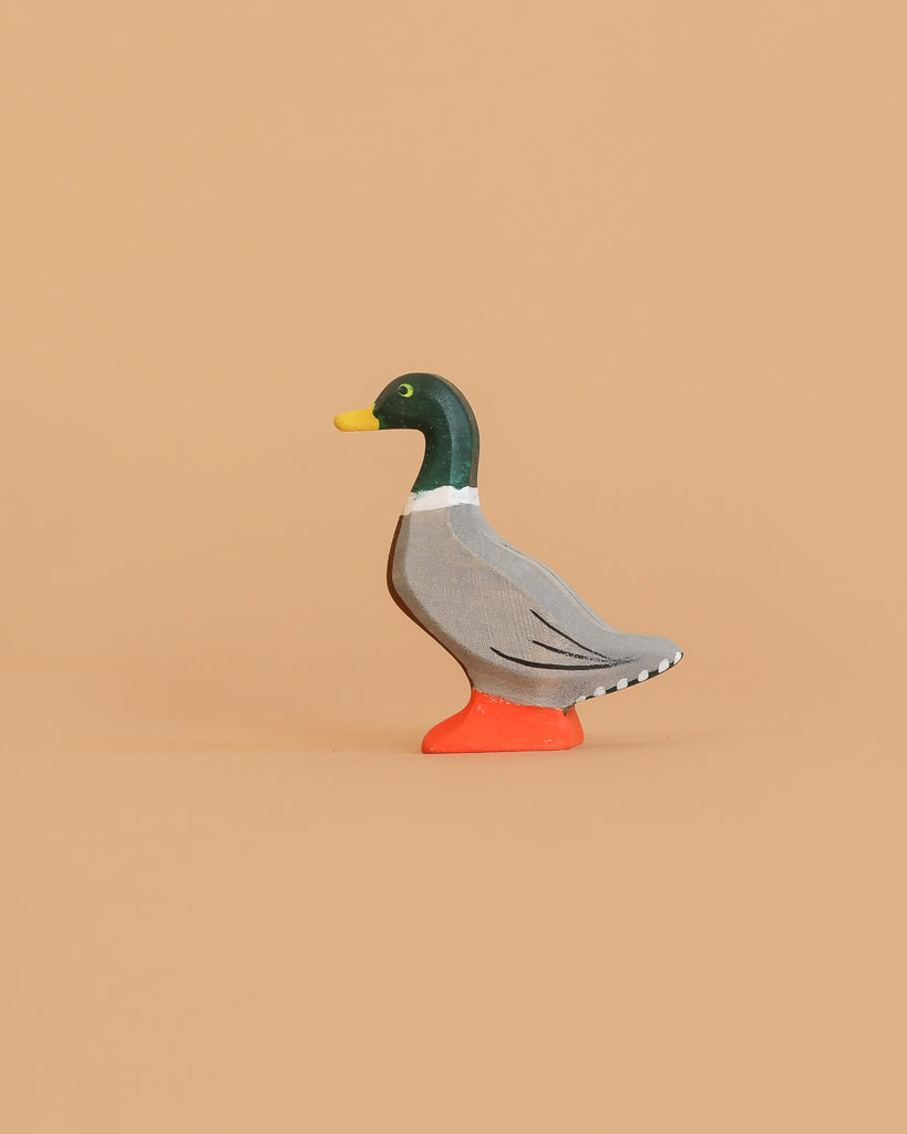 The Bumbu Handmade Wooden Mallard Duck, featuring a green head, yellow beak, gray body, and orange feet, stands on a plain beige background. This charming figurine is crafted from sustainable maple wood.