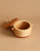 A round, light wooden container with a lid removed and placed to the side. The Handmade Wooden Pretend Pot has small handles on either side, and the interior is empty. Crafted from hardwoods like maple and beech, it sits against a uniform beige background.