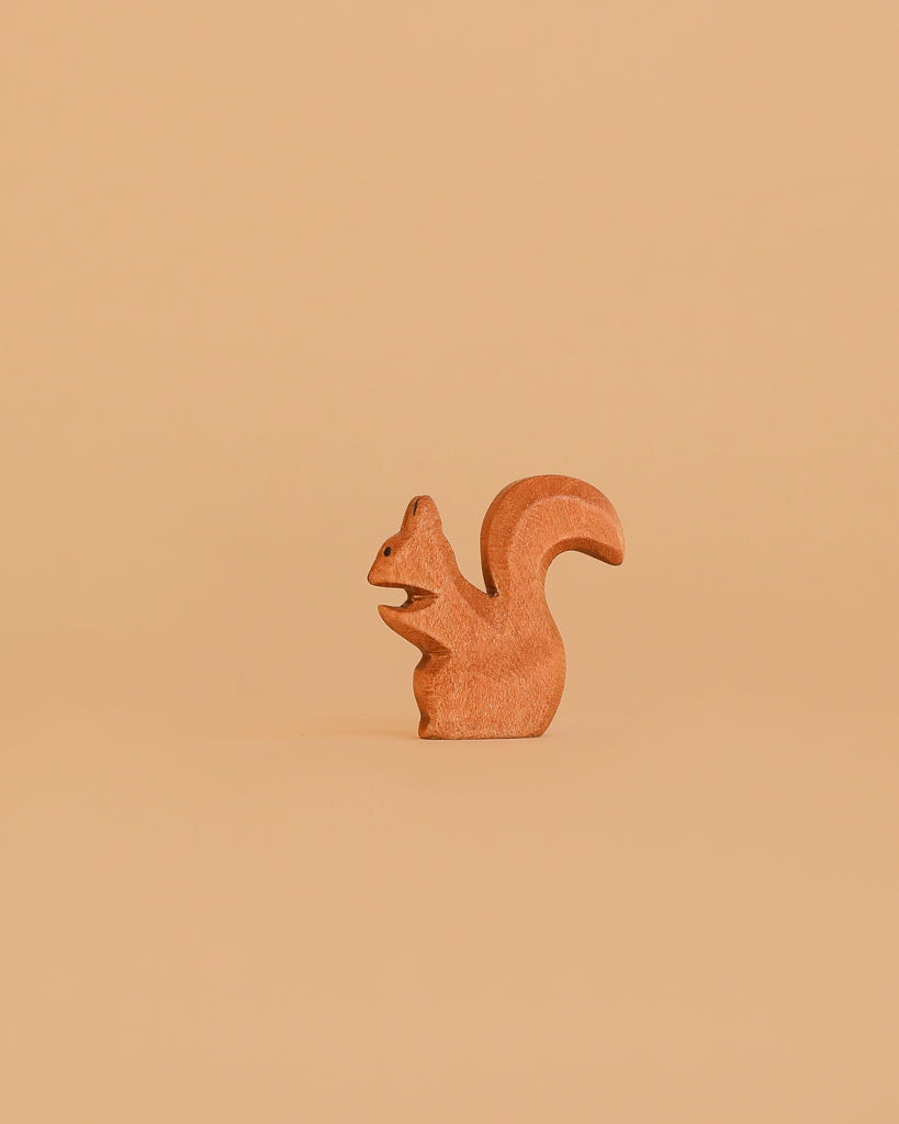 A small squirrel from the Bumbu Handmade Wooden Woodland Animals Set stands upright on a plain, light brown background, its tail curved upward and body facing left.