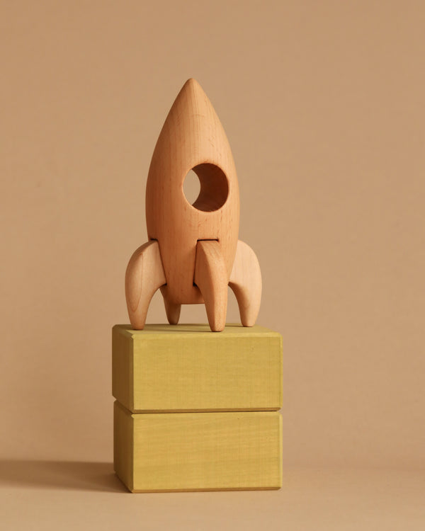 A Handmade Wooden Rocket with a circular window stands atop two stacked yellowish-green beech wood blocks. Finished with linseed oil, the background is a uniform light brown, creating a minimalist and neutral setting.