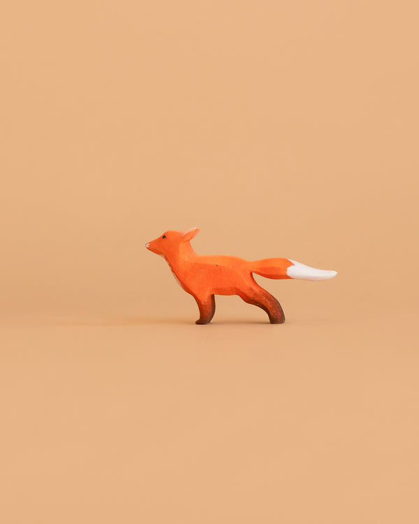 The Bumbu Handmade Wooden Fox Cub Curious, a small orange minimalist toy with a white-tipped tail, stands on a plain light brown background.