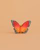 The Bumbu Handmade Wooden Orange Butterfly, featuring red and orange wings with black edges and white spots, stands upright against a plain beige background.