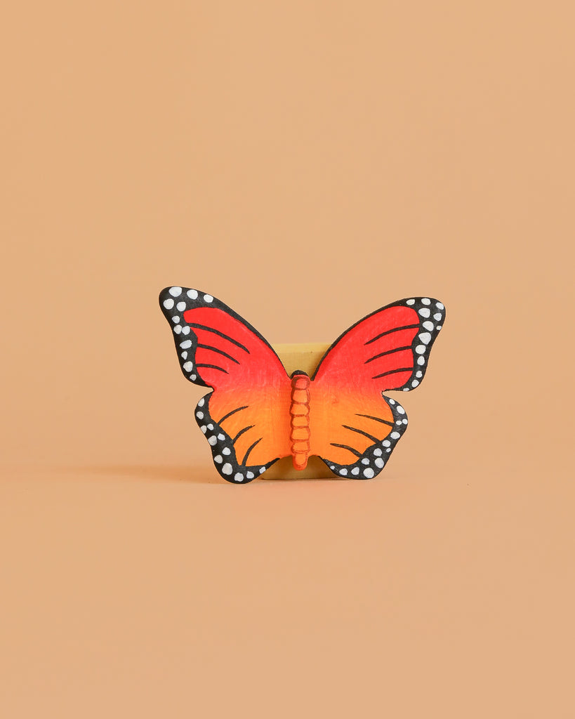 The Bumbu Handmade Wooden Orange Butterfly, featuring red and orange wings with black edges and white spots, stands upright against a plain beige background.