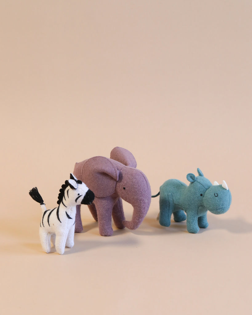 Three Olli Ella Holdie Folk Felt Safari Animals—a zebra, an elephant, and a rhinoceros—standing on a beige background. These handmade wool toys appear soft and are positioned in a staggered formation.