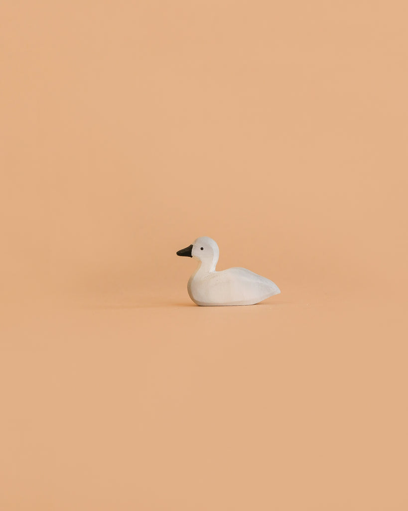 The Bumbu Handmade Wooden Baby Swan, a small white handcrafted wooden toy with a black beak, sits on a plain, light peach background.