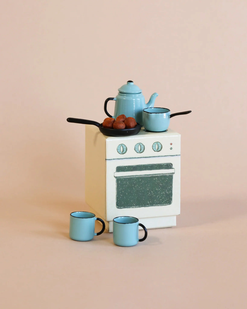 The Maileg Christmas Set features a charming miniature white oven with a light blue speckled door, adorned with small skillets and a kettle containing brown objects. Beside the oven, two matching blue mugs complete the cozy holiday scene against a plain beige background.