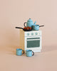 A miniature white stove with blue speckles is topped with small blue kitchenware and fake food, and is surrounded by three tiny blue cups on a plain beige background—an inviting scene reminiscent of cozy evenings spent in the Maileg Winter Chalet Set.