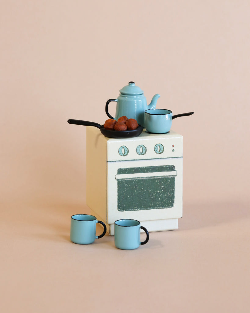 A miniature white stove with blue speckles is topped with small blue kitchenware and fake food, and is surrounded by three tiny blue cups on a plain beige background—an inviting scene reminiscent of cozy evenings spent in the Maileg Winter Chalet Set.
