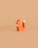 A small, orange minimalist fox with a white-tipped tail and chest sits on beige—an adorable addition to any collection, like the Bumbu Handmade Wooden Woodland Animals Set.
