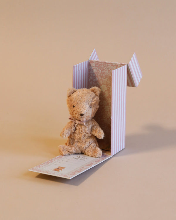 A small, plush Maileg My First Teddy, Powder is sitting inside an open striped cardboard box with a vintage look and floral inner lining. The box is slightly tilted, its lid open, revealing a decorative card with a teddy bear image in front of it. The background is a neutral beige, perfect for welcoming a newborn baby.