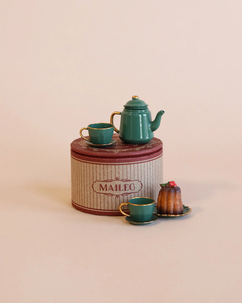 The Maileg Ski Lounge Set offers a charming vintage-style teal tea set, complete with two cups and a teapot, elegantly displayed on a decorative tin. Accompanying the tea set is a small Bundt cake topped with a cherry, placed on a matching plate—ideal for enjoying an afternoon with the big brother ski mouse.