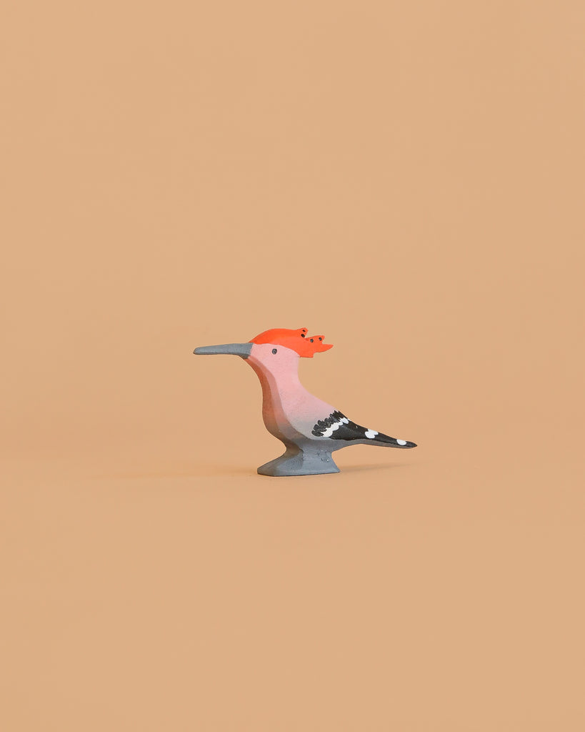 The Bumbu Handmade Wooden Hoopoe Bird is a small, handcrafted wooden figurine featuring an orange crest, pink body, gray beak, and black-and-white striped tail set against a plain beige background.