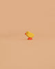 The Bumbu Handmade Wooden Chick, a small, bright yellow figurine with a red beak and orange feet, is crafted from sustainable wood and displayed against a plain light tan background.