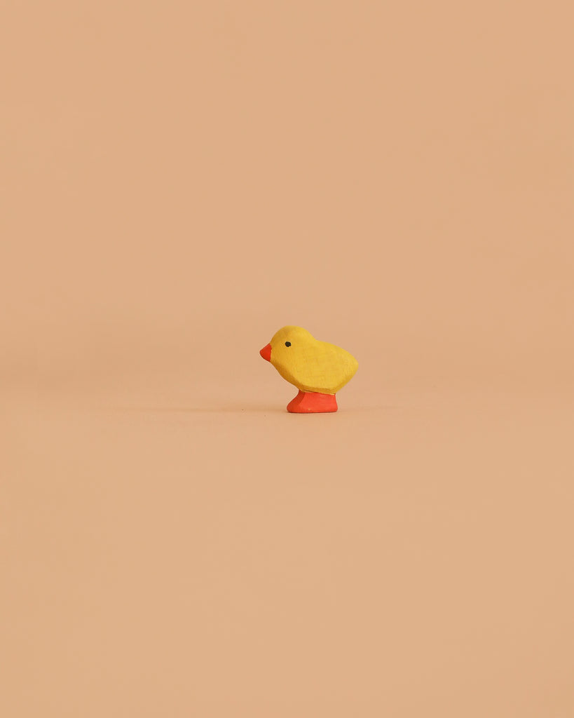 The Bumbu Handmade Wooden Chick, a small, bright yellow figurine with a red beak and orange feet, is crafted from sustainable wood and displayed against a plain light tan background.