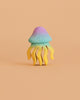 The Bumbu Handmade Wooden Jellyfish, a small pastel sculpture with a blue-purple dome and yellow wavy tentacles, creates an enchanting eco-friendly toy when displayed against a plain peach background.