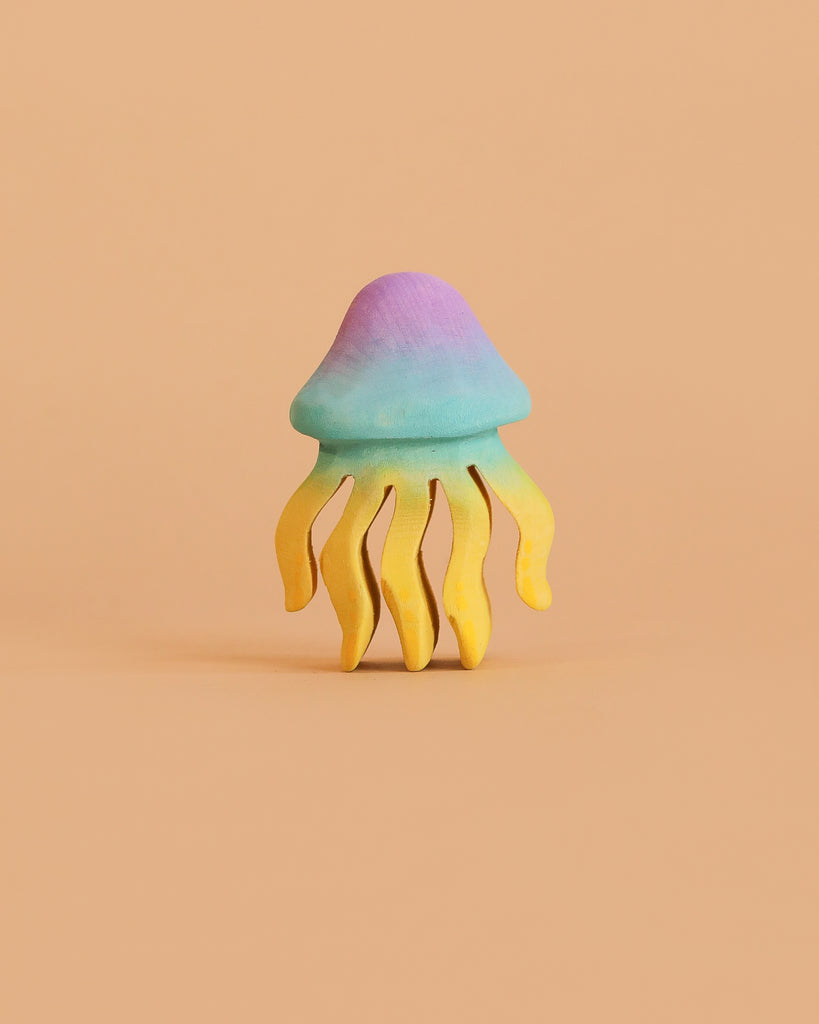 The Bumbu Handmade Wooden Jellyfish, a small pastel sculpture with a blue-purple dome and yellow wavy tentacles, creates an enchanting eco-friendly toy when displayed against a plain peach background.