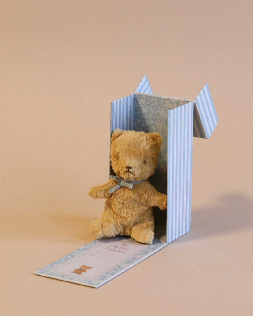 A small, light brown **Maileg My First Teddy, Sand** with a blue ribbon around its neck sits inside an open blue and white striped gift box on a tan background. The lid of the box is open, and there is a card with a bear illustration in front of the box, making it a perfect new born baby gift.