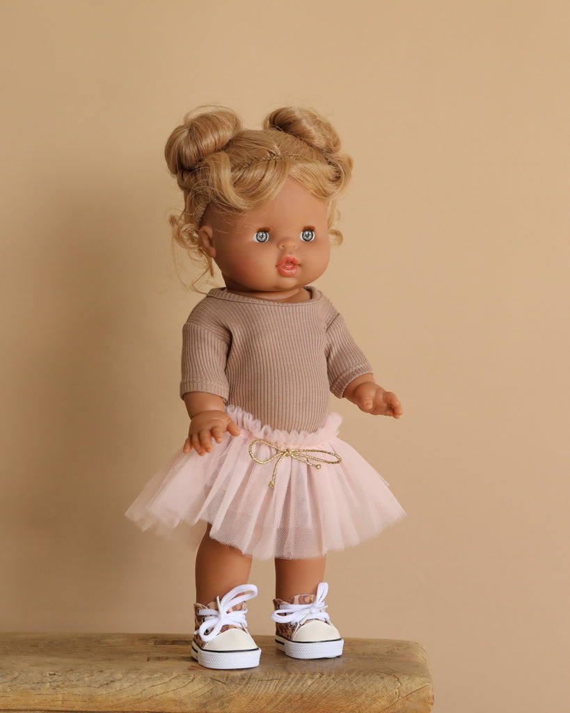 The Minikane Standing Doll (14") - Louisa in Brown Tutu features blonde hair in buns and stands on a wooden surface against a beige backdrop. She wears a brown long-sleeve top, pink tutu skirt with a bow, white sneakers with blue laces, and has a natural vanilla scent.