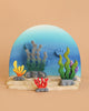 The Bumbu Handmade Wooden Deep Ocean Water Set features colorful marine toys with seaweed and coral designs on a beige base, set against a blue gradient backdrop. These vibrant pieces encourage imaginative play with green, yellow, gray, and red plant shapes.
