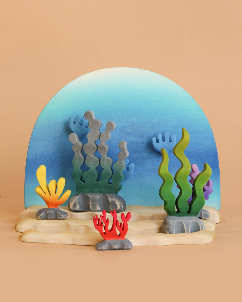 The Bumbu Handmade Wooden Deep Ocean Water Set features colorful marine toys with seaweed and coral designs on a beige base, set against a blue gradient backdrop. These vibrant pieces encourage imaginative play with green, yellow, gray, and red plant shapes.