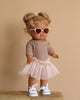 The Minikane Standing Doll (14") - Louisa in a brown tutu, styled with pink heart-shaped sunglasses and white sneakers, stands on a wooden platform. The neutral background enhances her playful charm and natural vanilla scent.