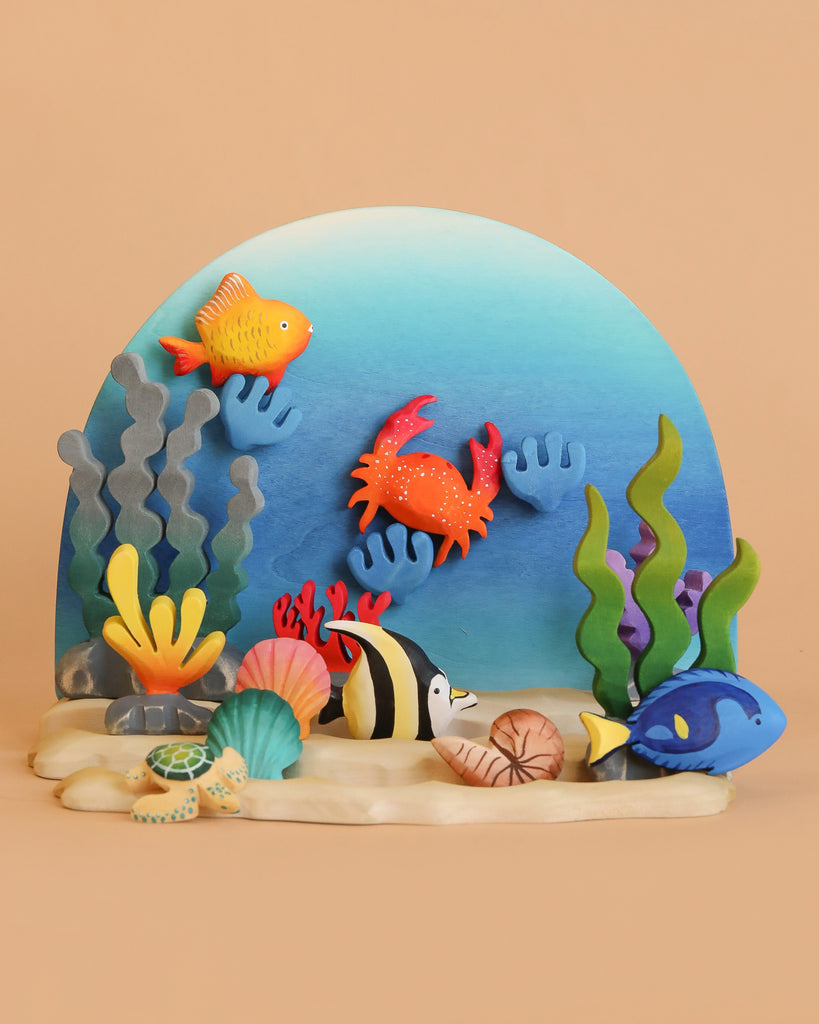 The Bumbu Handmade Wooden Ocean Set, featuring crab and turtle figures, is displayed in an underwater scene with a blue background, sandy foreground, and peach backdrop alongside colorful clay sea creatures.