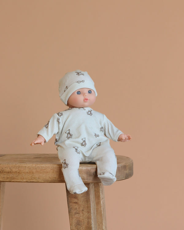 The Minikane Soft Body Doll (11") – Claire With Clothing, dressed in a matching cream outfit and bear-print hat, sits on a wooden stool against a plain beige background.