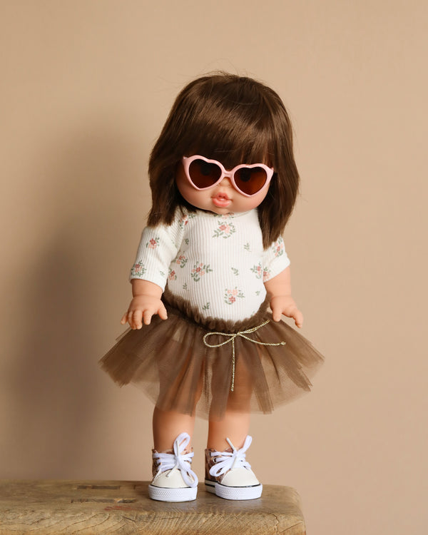 A Minikane Standing Doll (14") - Zoelie In Pink Tutu is posed on a wooden stool against a beige background, donning heart-shaped sunglasses, a floral long-sleeve shirt, brown tulle skirt, white shoes with purple laces, and gives off a hint of natural vanilla scent. Ships in one week.