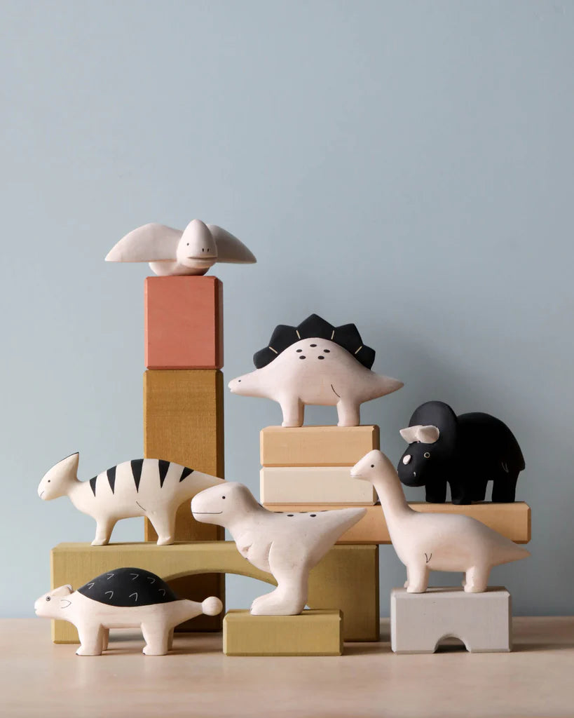 A collection of hand-painted wooden Stegosaurus figurines, artistically arranged on and around pastel-colored blocks against a pale blue background.